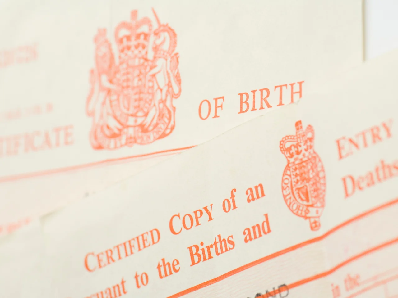 A close-up of official UK birth certificate documents, featuring a red royal crest and partially visible text reading CERTIFIED COPY OF AN ENTRY, with other text and emblems faintly in the background.