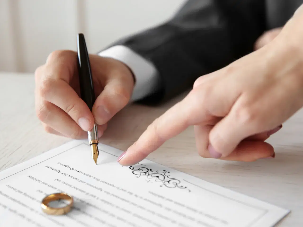 A person in a suit signs a document with a fountain pen while another hand points to a section. Two gold wedding rings rest on the table beside the paper.