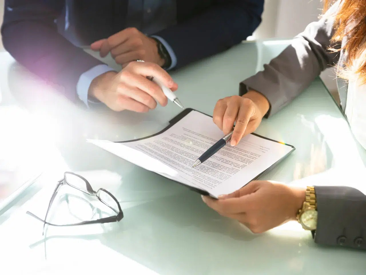 Two people in business attire discuss a document with printed text on a clipboard, pointing with pens. A pair of eyeglasses rests on the glass table beside them, with sunlight streaming in from the left.