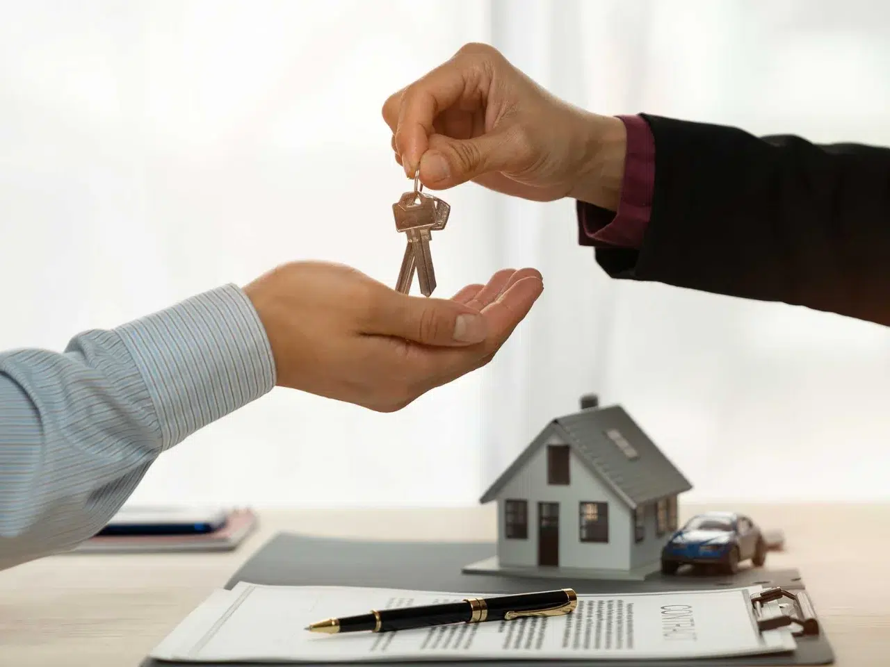 A person hands over a set of keys to another person. In the background, there is a small model house and a toy car on a table with a contract and a pen, suggesting a real estate transaction.