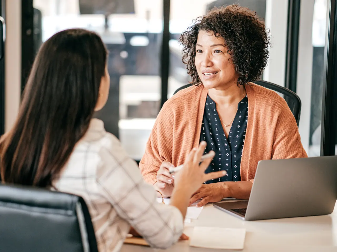 Two women sit at a desk in an office, having a conversation. One woman is facing the camera, smiling, with a laptop open in front of her; the other has her back to the camera and gestures with her hands.