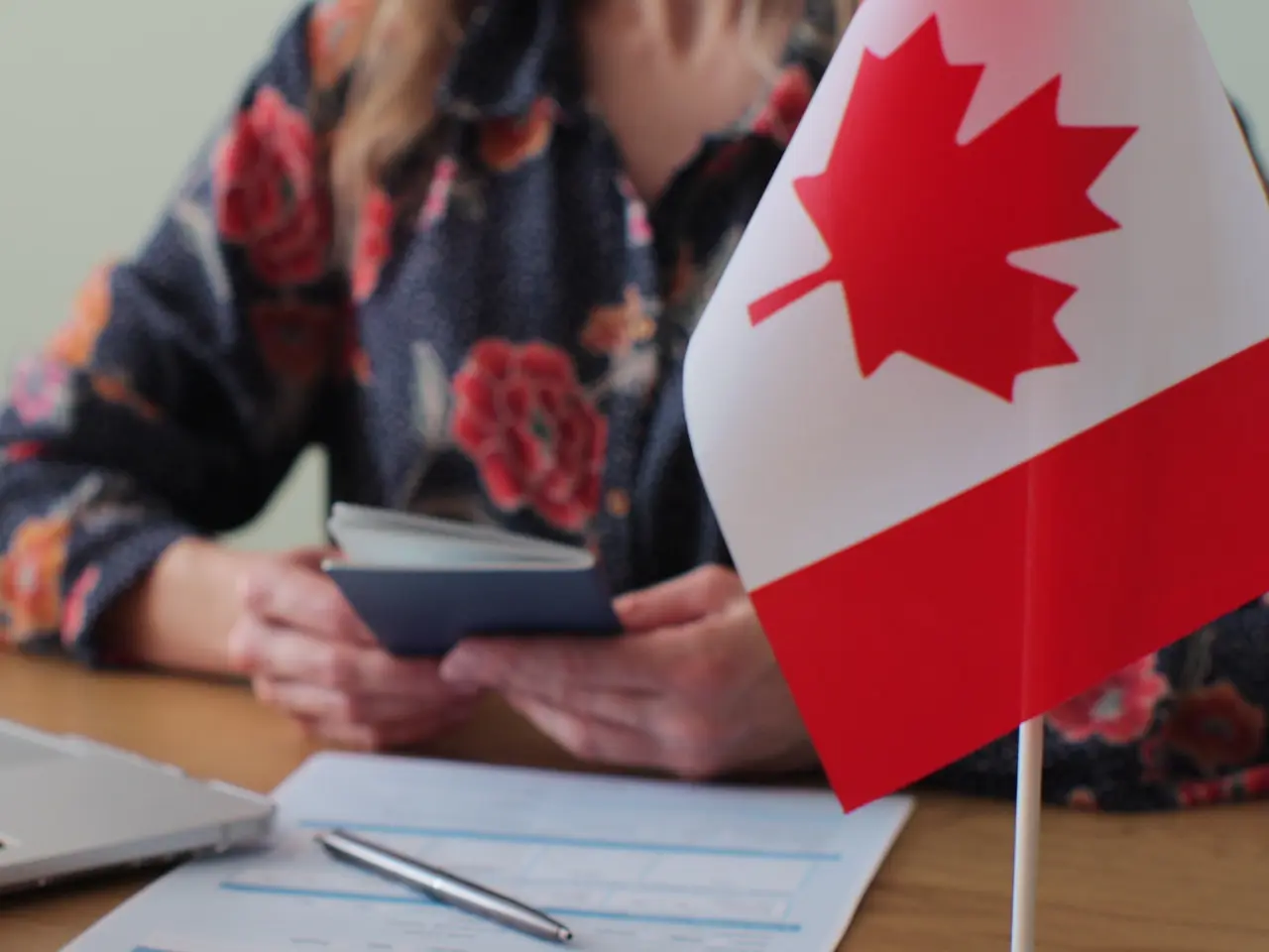 A person holding a passport sits at a table with immigration forms, a pen, and a small Canadian flag in the foreground.
