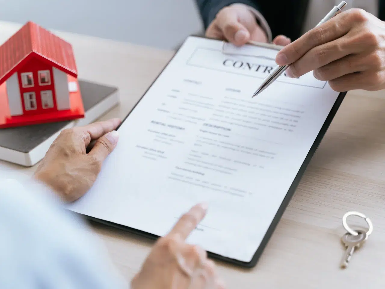 Two people review and discuss a contract on a clipboard at a desk. A small model house, a book, and a set of keys are nearby, suggesting a real estate or property agreement.