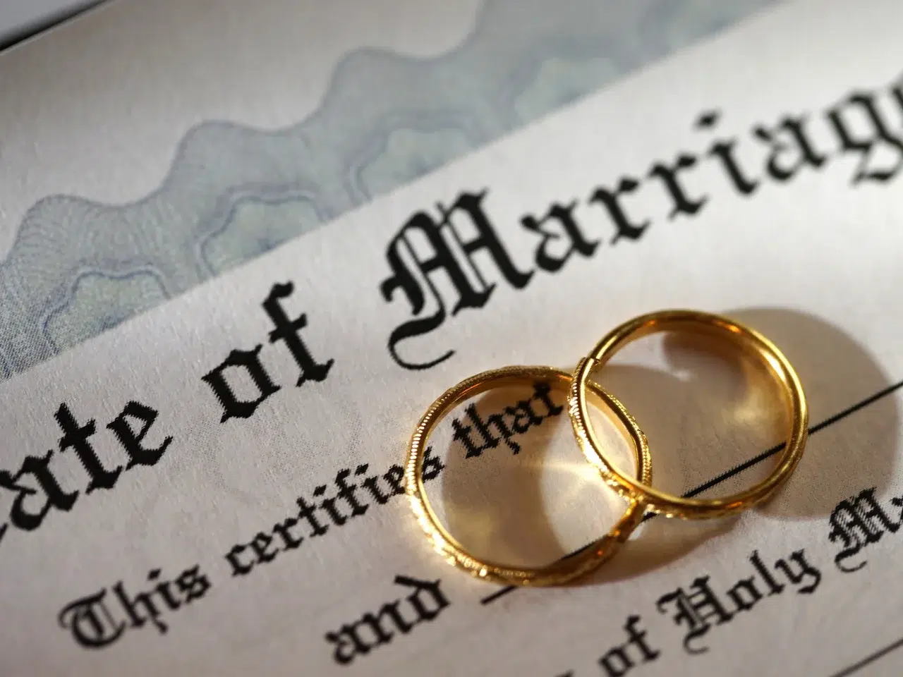 Two gold wedding rings rest on top of a marriage certificate, partially covering the text. The focus is on the rings and the word Marriage in large, decorative font.