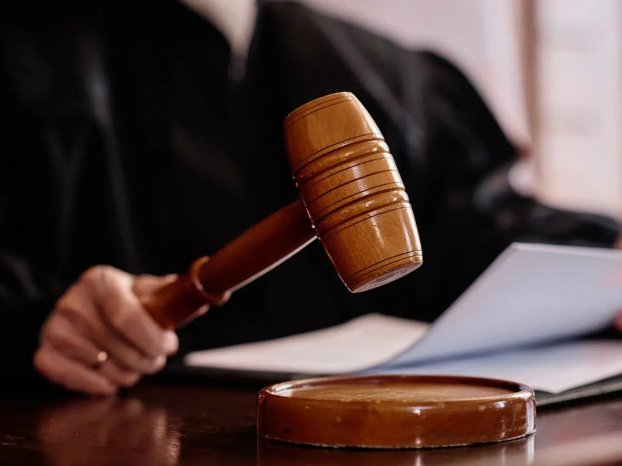 A judge holding a wooden gavel above a sound block, with paperwork in the other hand, wearing a black robe, seated at a desk in a courtroom.