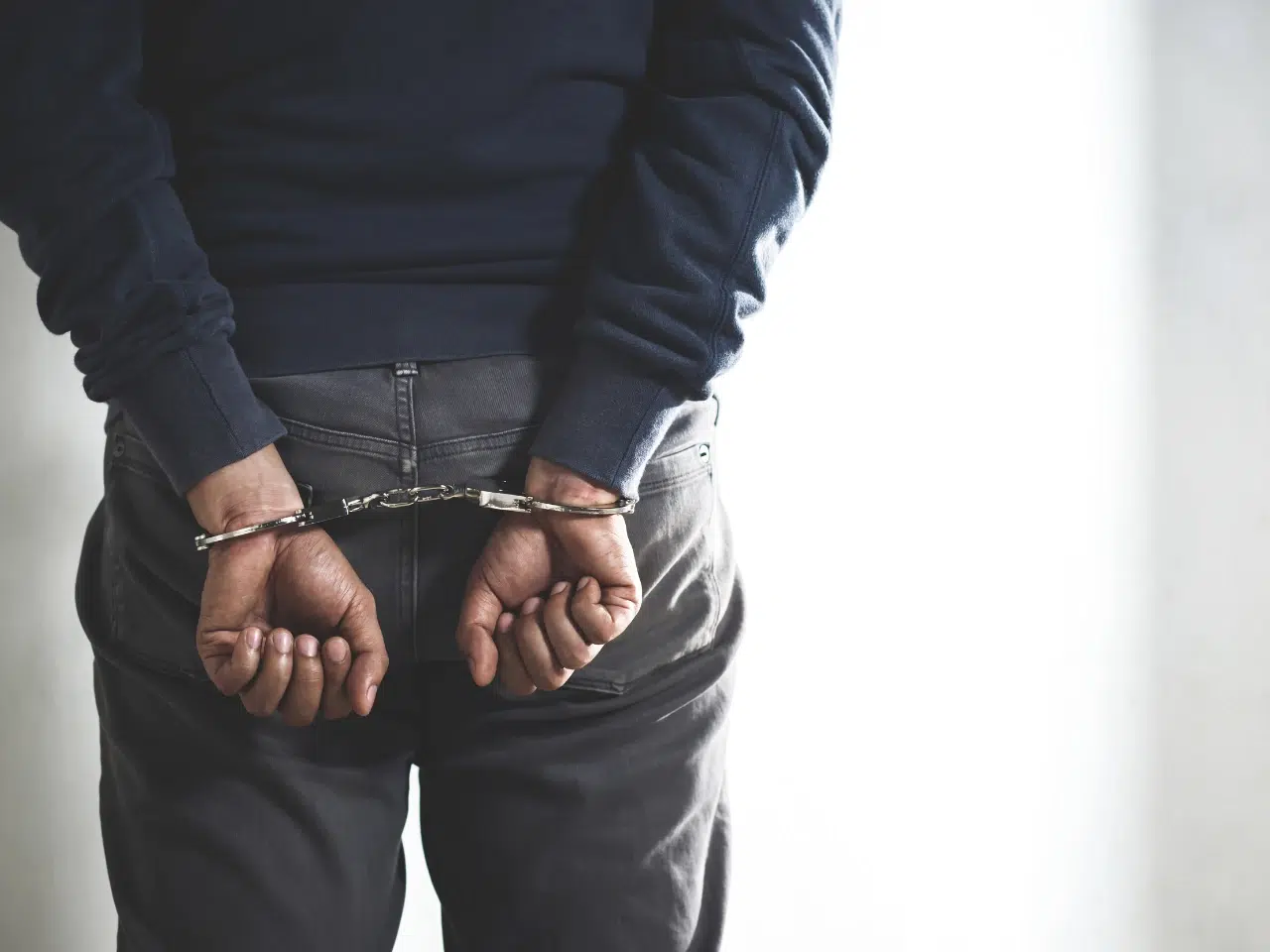 A person wearing dark clothing is shown from behind with their hands cuffed behind their back, suggesting they have been arrested or detained. The background is plain and light-colored.