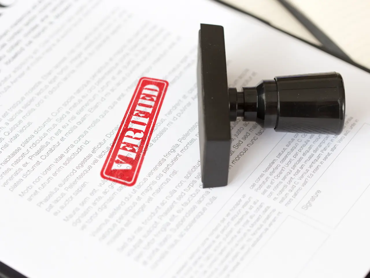 A document with a large black stamp resting on it, displaying a bold red VERIFIED mark in the center of the page. The text of the document is mostly blurred and unreadable.