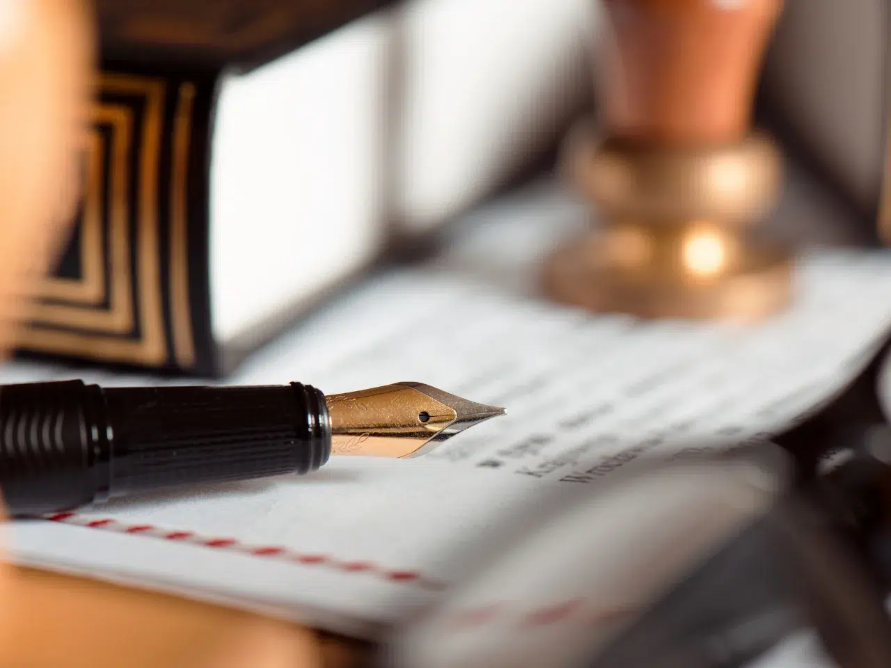 A close-up of a fountain pen resting on a document, with a book and a wooden stamp in the blurred background, suggesting a legal or official setting.