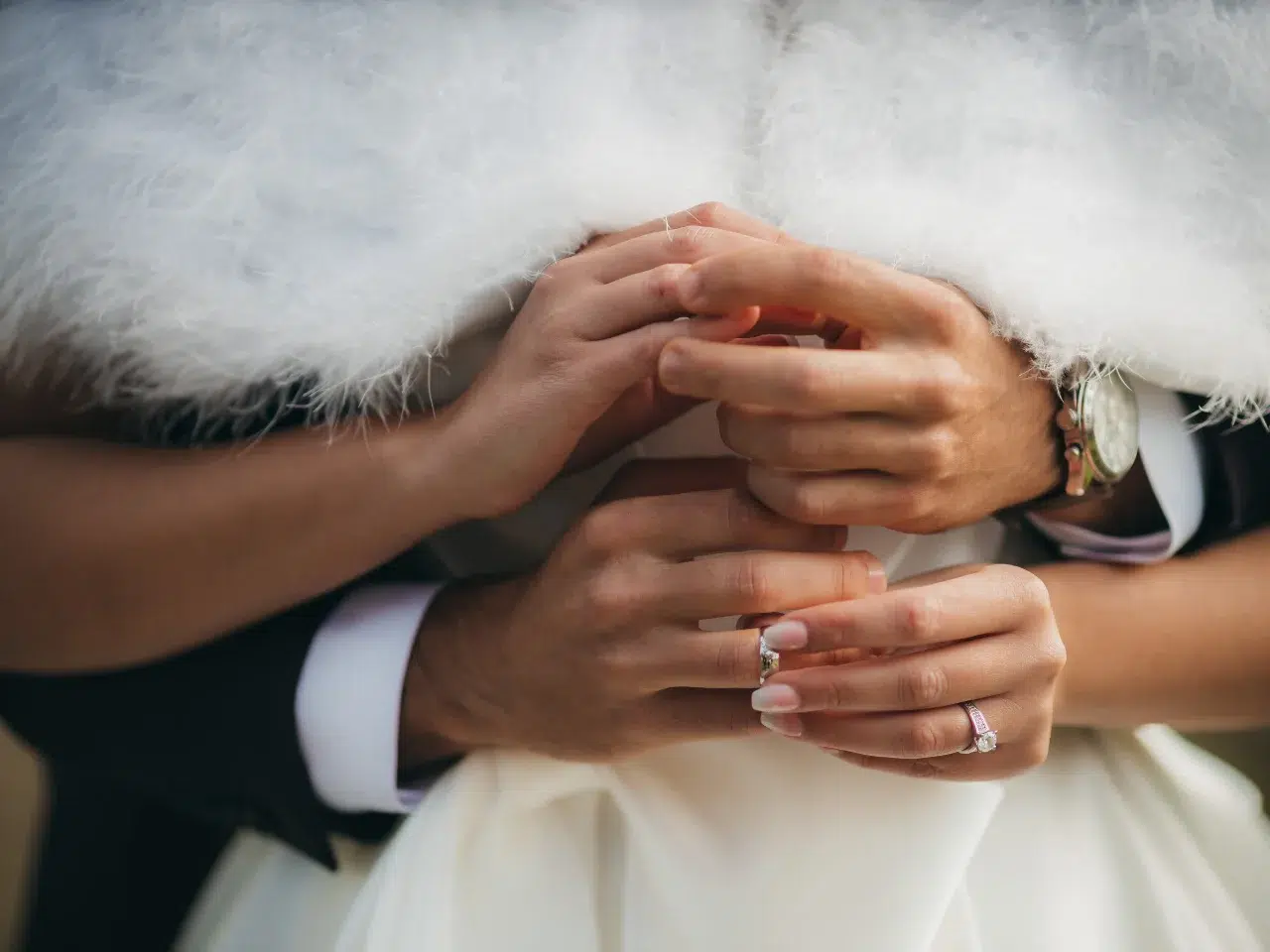 Close-up of two people embracing, showing their hands gently holding each other. They are dressed formally, and both wear rings, suggesting a romantic or wedding moment. One person has a white fluffy shawl.