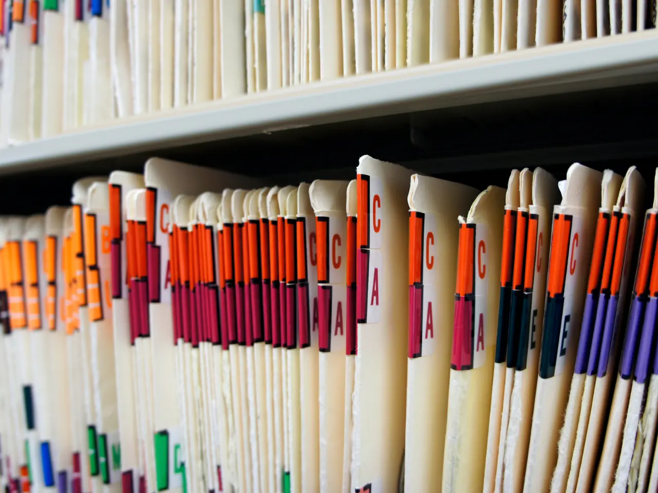 Close-up of a row of file folders with colored lettered tabs, arranged upright on a shelf, suggesting an organized filing or record-keeping system. More files are visible on the shelf above.