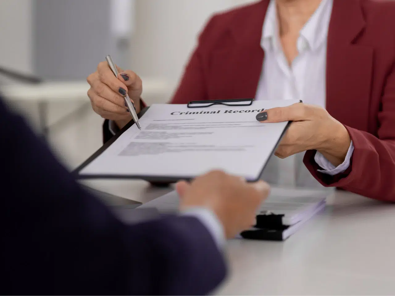 Two people sit at a desk; one hands a clipboard labeled Criminal Record to the other. Both are wearing business attire, and paperwork is visible on the table.