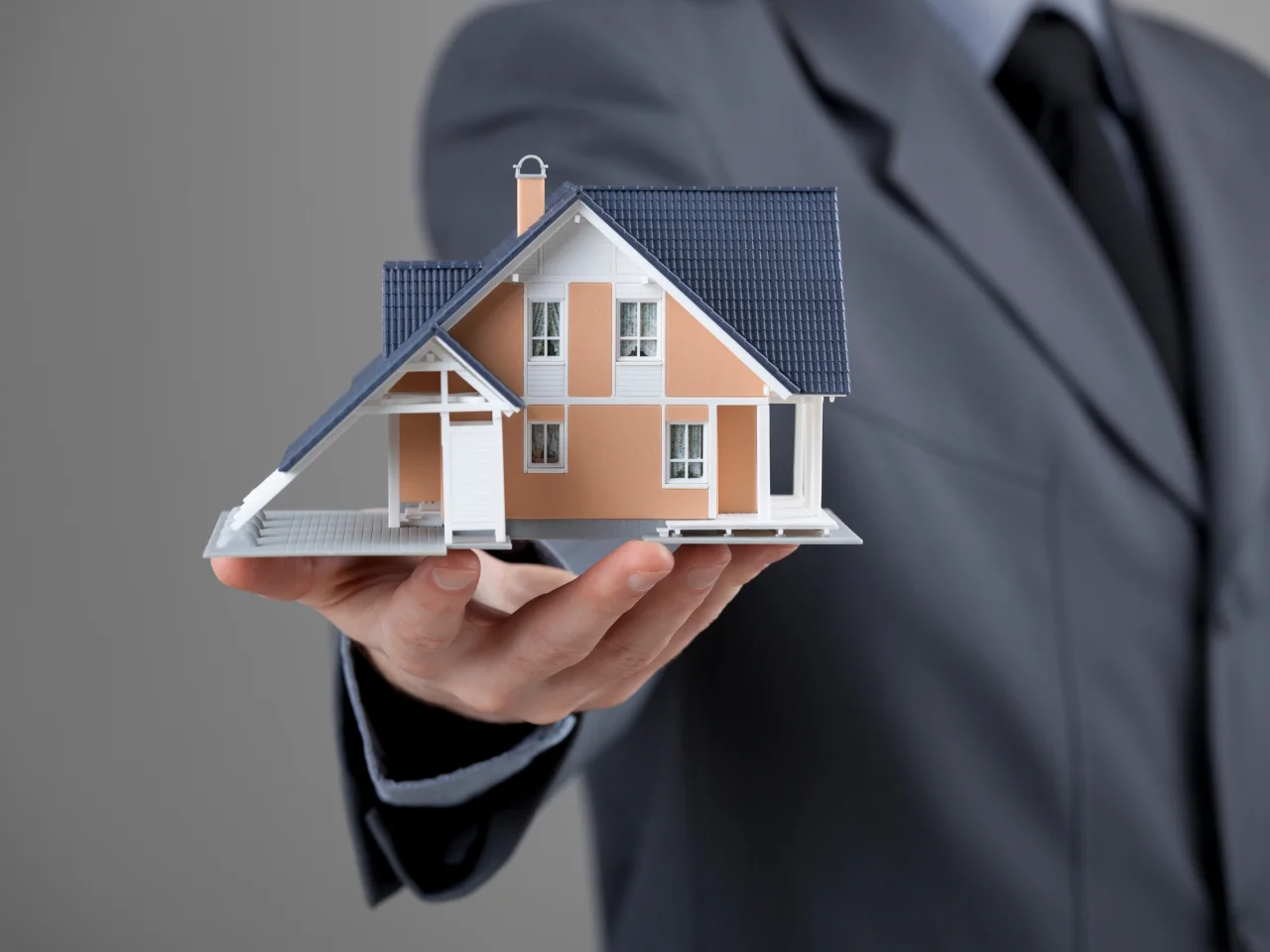 A person in a suit holds a detailed model of a house with blue roof tiles and beige walls, symbolizing real estate, home ownership, or property investment. The background is plain and gray.