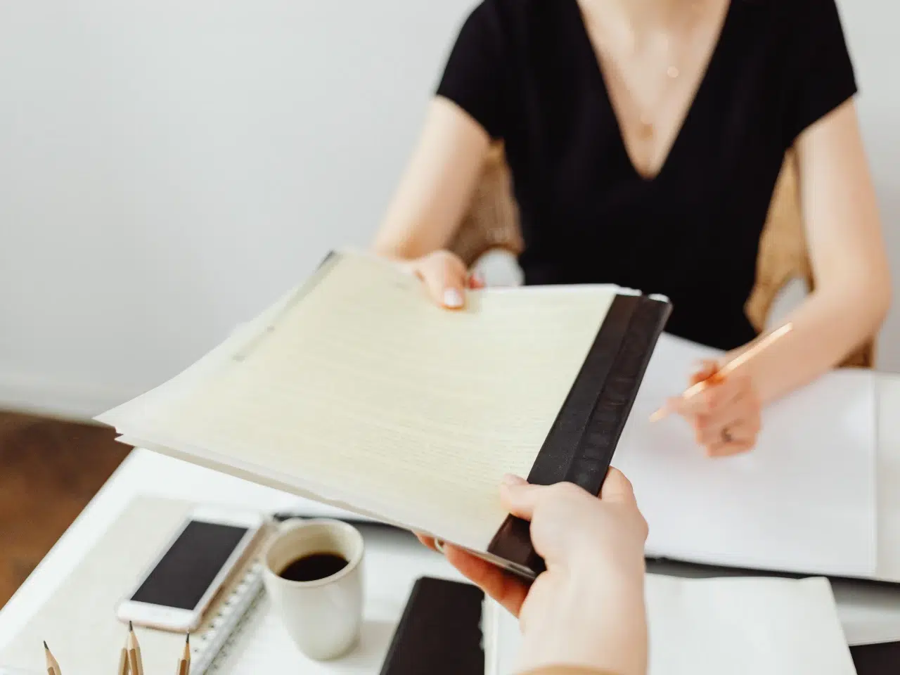 A person hands a document to a woman sitting at a desk with a notebook, pen, smartphone, and a cup of coffee, suggesting a professional or office setting.