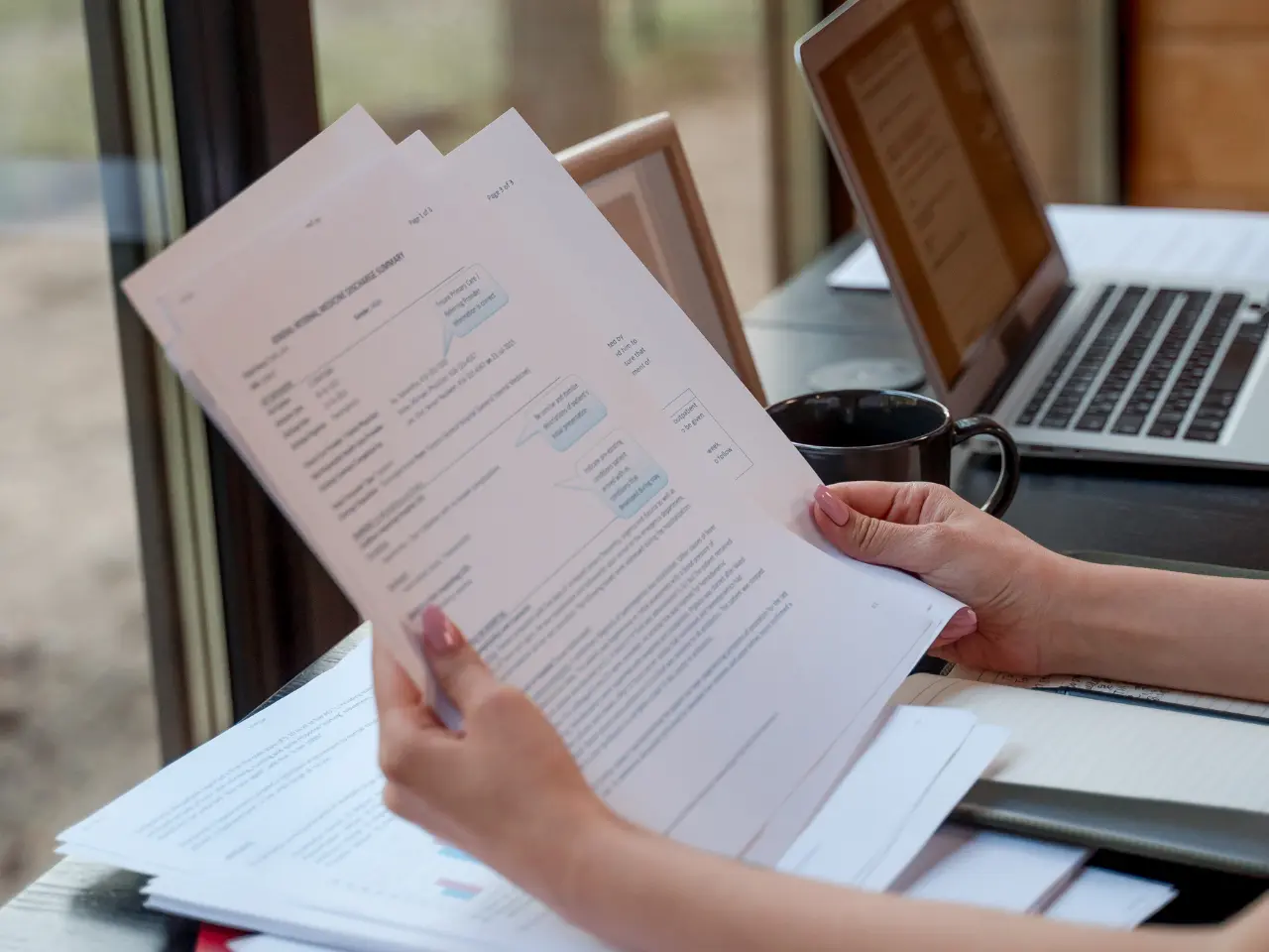 A person holding and reading printed documents at a desk with a laptop, papers, and a coffee mug, near a window with an outdoor view.