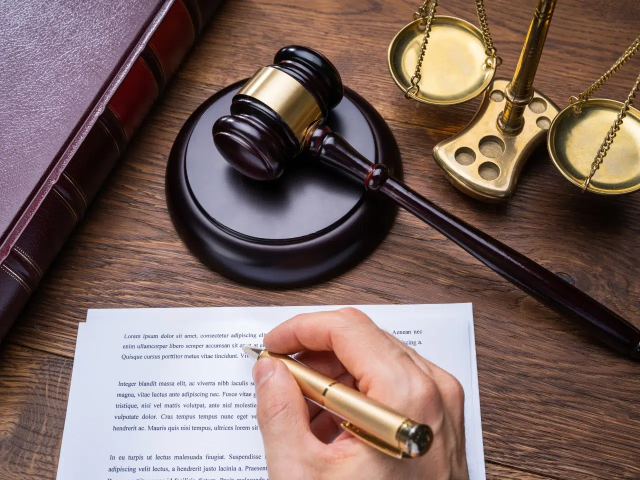 A hand holding a pen signs a document on a wooden desk beside a judges gavel, golden scales of justice, and a large brown legal book.