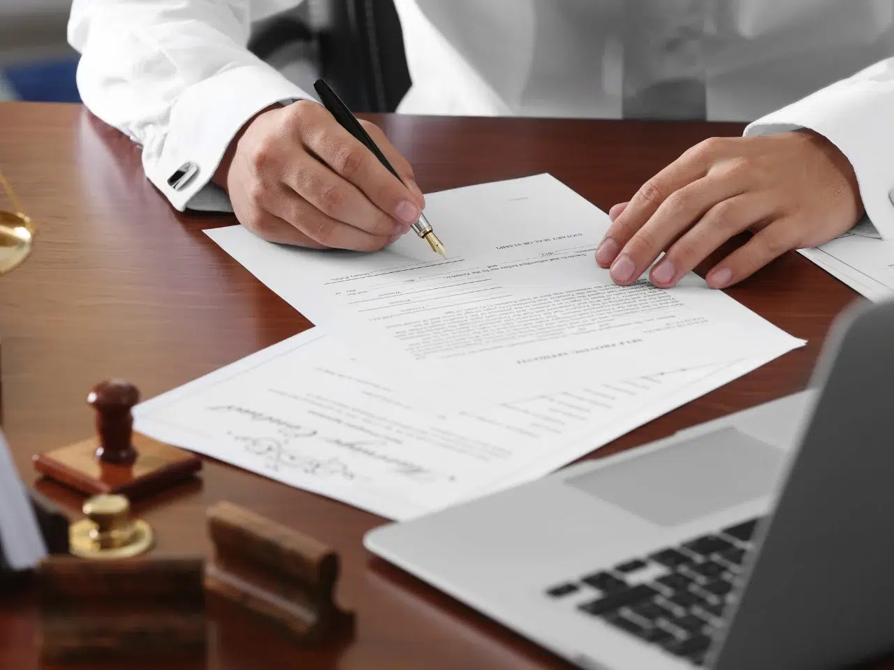 A person in a white shirt signs a document at a wooden desk, with a laptop, a wax seal stamp, and other paperwork nearby.