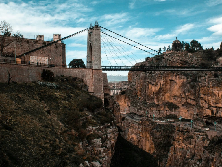 A suspension bridge spans a deep rocky gorge under a partly cloudy sky, connecting stone buildings on either side amid rugged, green-accented cliffs.
