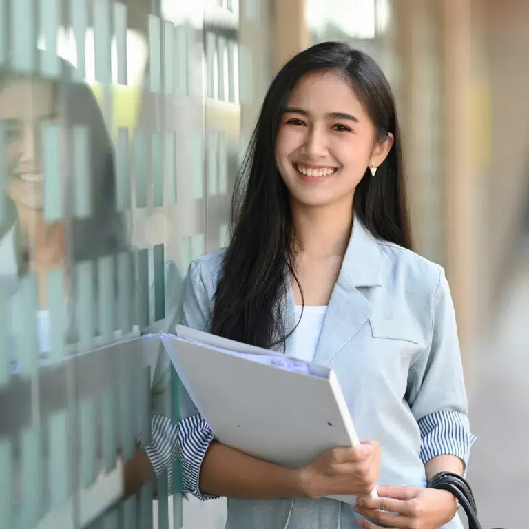 A young woman with long dark hair, wearing a light blazer, smiles while holding folders and standing next to a glass wall with reflections.