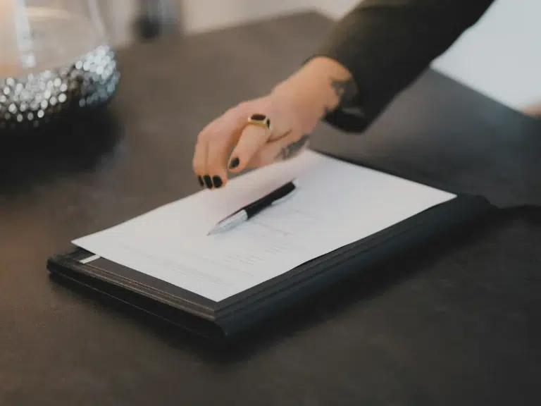 A person with black nail polish and a ring points to a document on a clipboard, with a pen resting on top, on a dark table.