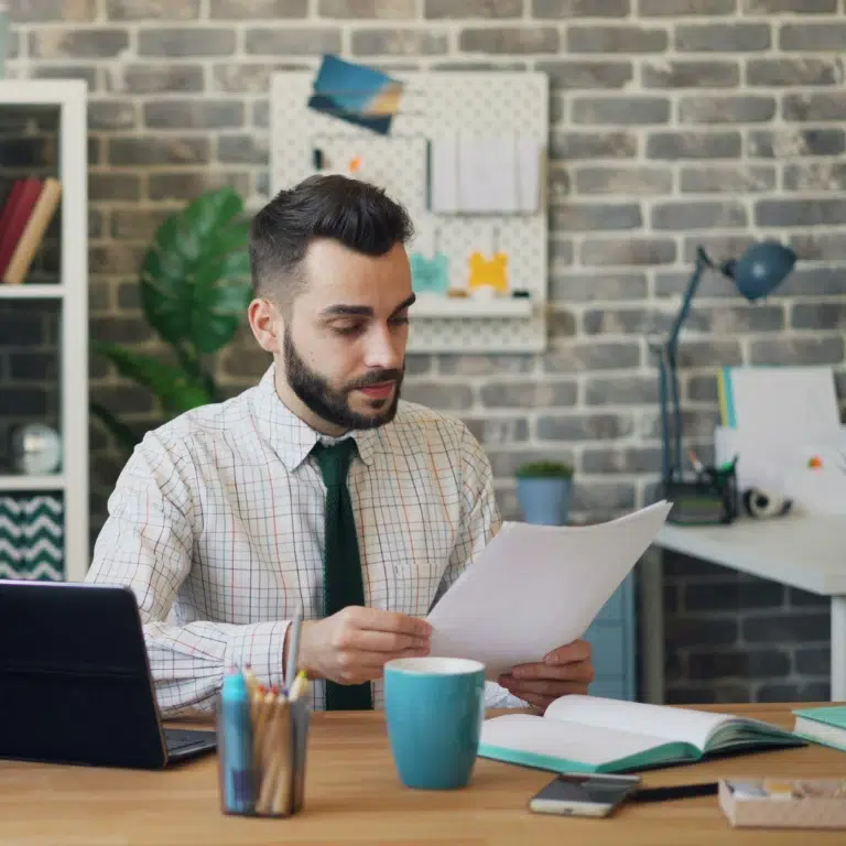 A man in a checked shirt and tie sits at a desk with papers, a coffee mug, a tablet, and office supplies, working in a modern office with a brick wall and organizational boards in the background.