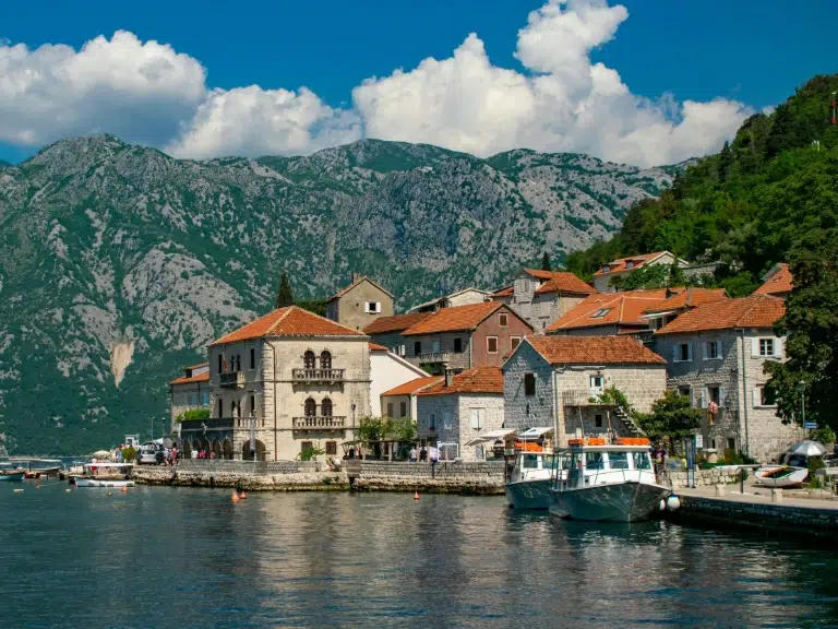 A picturesque coastal village with stone houses and red roofs sits beside calm water, backed by green mountains under a partly cloudy sky; a white boat is docked at the waterfront.