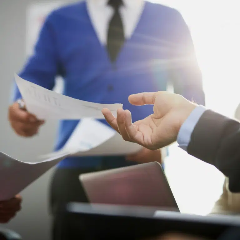Three people in business attire exchanging documents during a meeting, with one person handing a paper to another. Sunlight streams in from the background, and an open laptop is visible in the foreground.