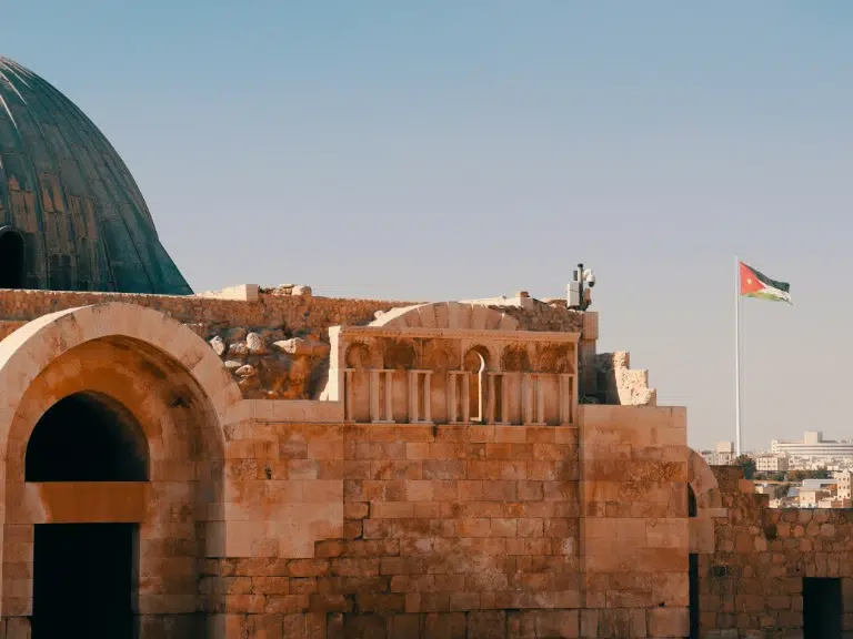 Ancient stone structure with an arched doorway and dome, set against a clear sky. A Jordanian flag waves in the distance, and city buildings are visible on the horizon.