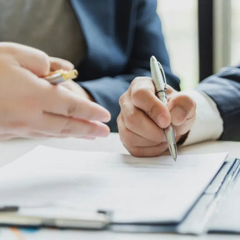 Two people in business attire are discussing a document on a desk, possibly finalizing paperwork for rcmp accredited fingerprinting companies. One person is holding a pen to sign, while the other gestures with their hand.