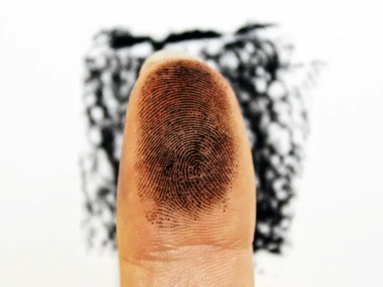 A close-up of a fingertip covered in black ink, showing detailed fingerprint ridges. In the background, a fingerprint impression is visible on a white surface, commonly used by RCMP accredited fingerprinting companies.