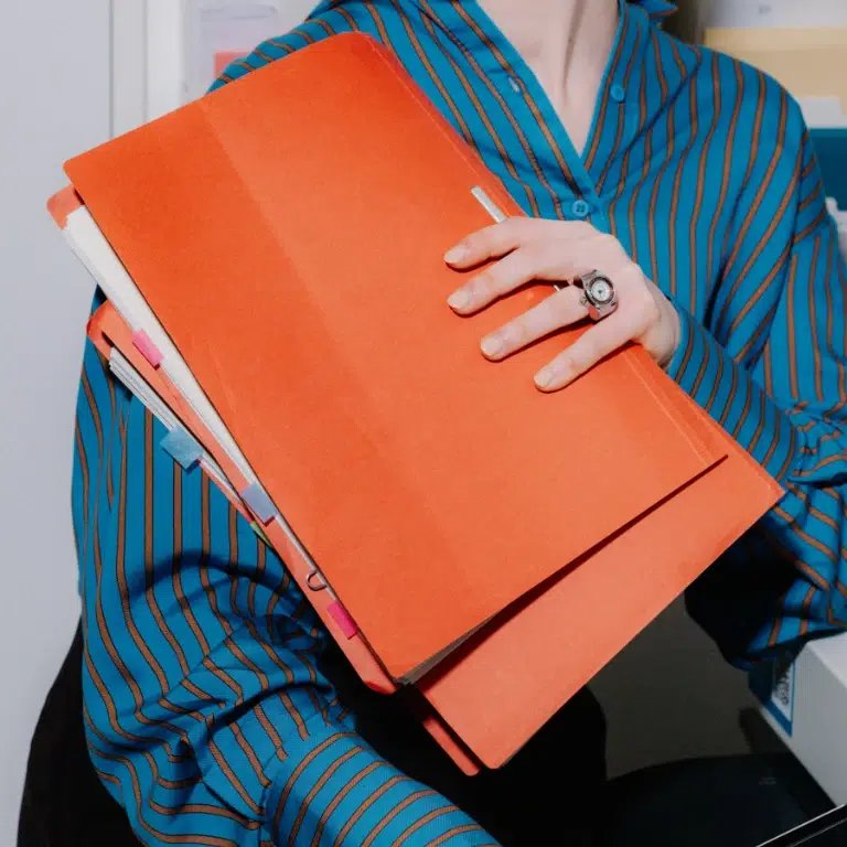 A person wearing a blue and black striped shirt holds several large orange file folders filled with papers and documents, some with colorful tabs visible.