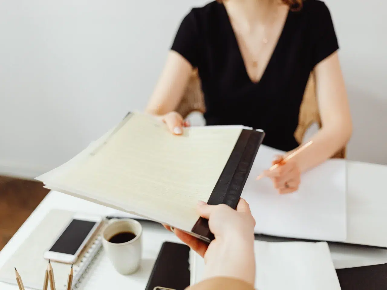 A person hands a clipboard with papers to a woman in a black dress sitting at a desk with a notebook, pen, smartphone, and a cup of coffee. The setting appears to be an office or meeting.