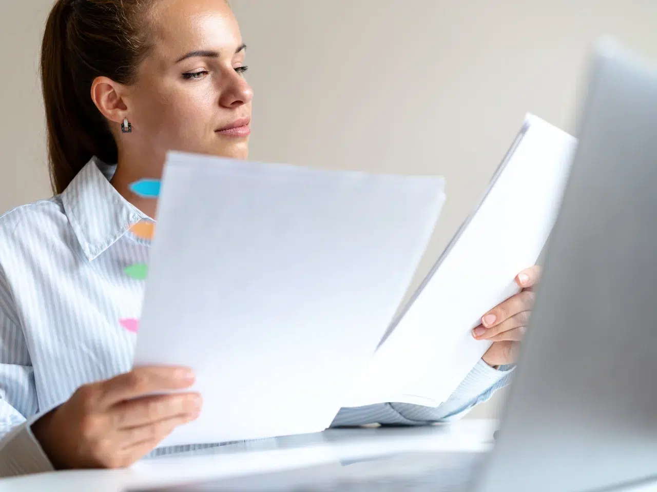 A woman sits at a desk, holding and reviewing documents in her hands. She appears focused, with colorful sticky notes visible next to her and a laptop open in front of her.