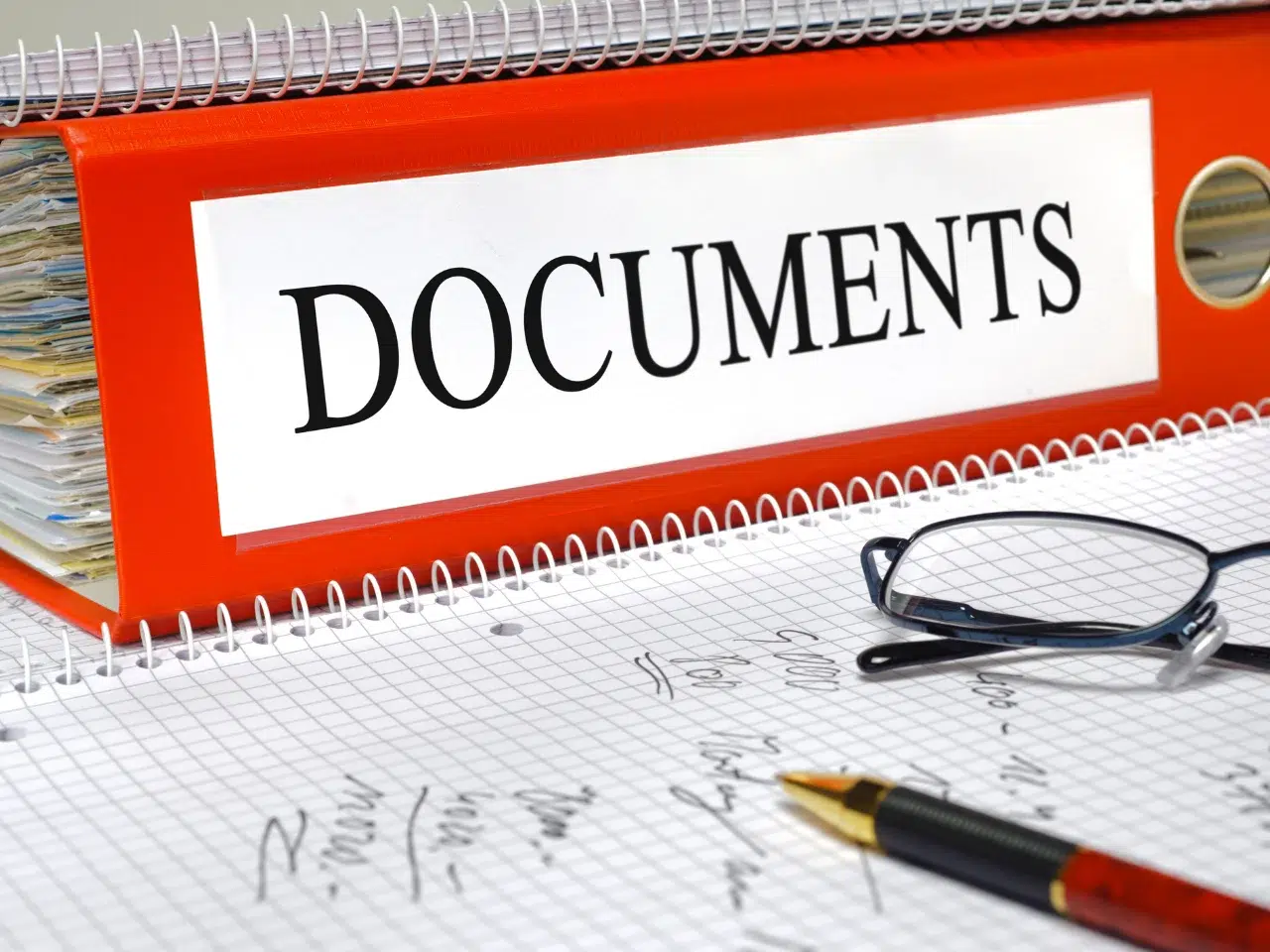 A red binder labeled DOCUMENTS sits on a table with a pair of glasses, a black and gold pen, and a notebook filled with handwritten notes in the foreground.
