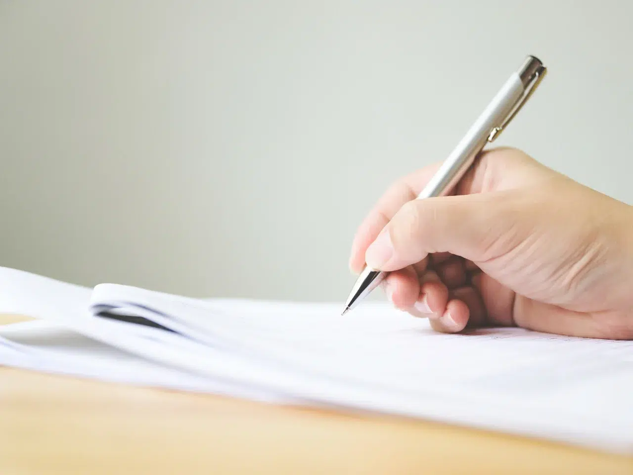 A person’s hand holding a silver pen is writing on a stack of white paper on a light wooden desk, with a plain light background.