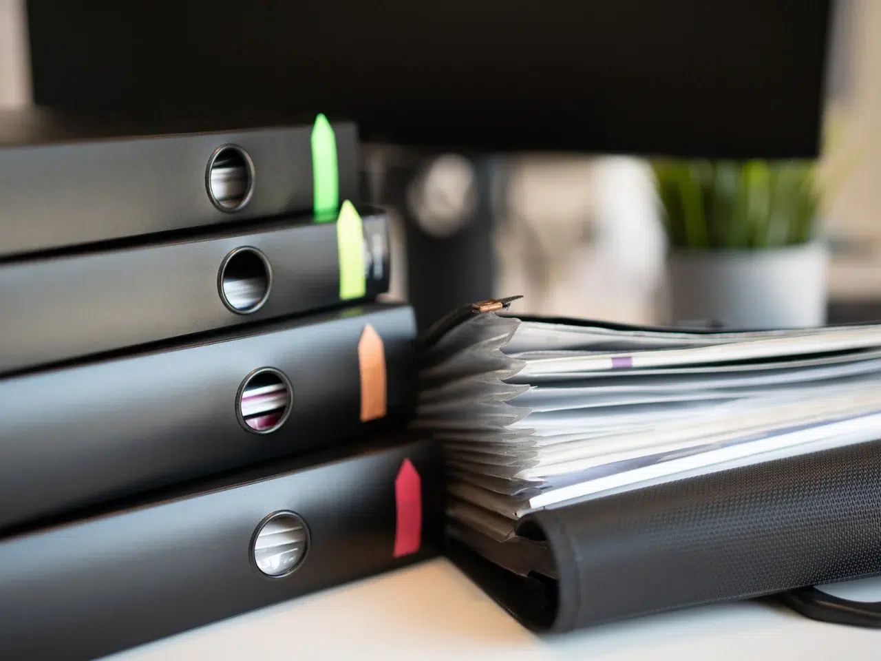A close-up of stacked black office binders with colored labels, next to an open binder filled with papers. A blurred computer monitor and a potted plant are visible in the background.