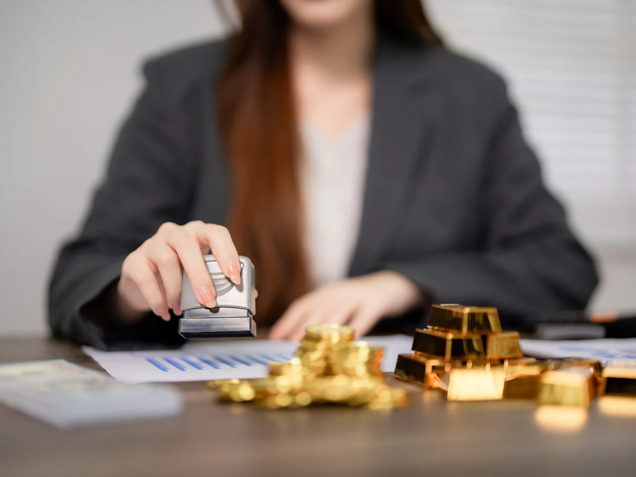 A woman in a blazer stamps a document with charts, while stacks of gold bars and coins are on the desk in front of her.