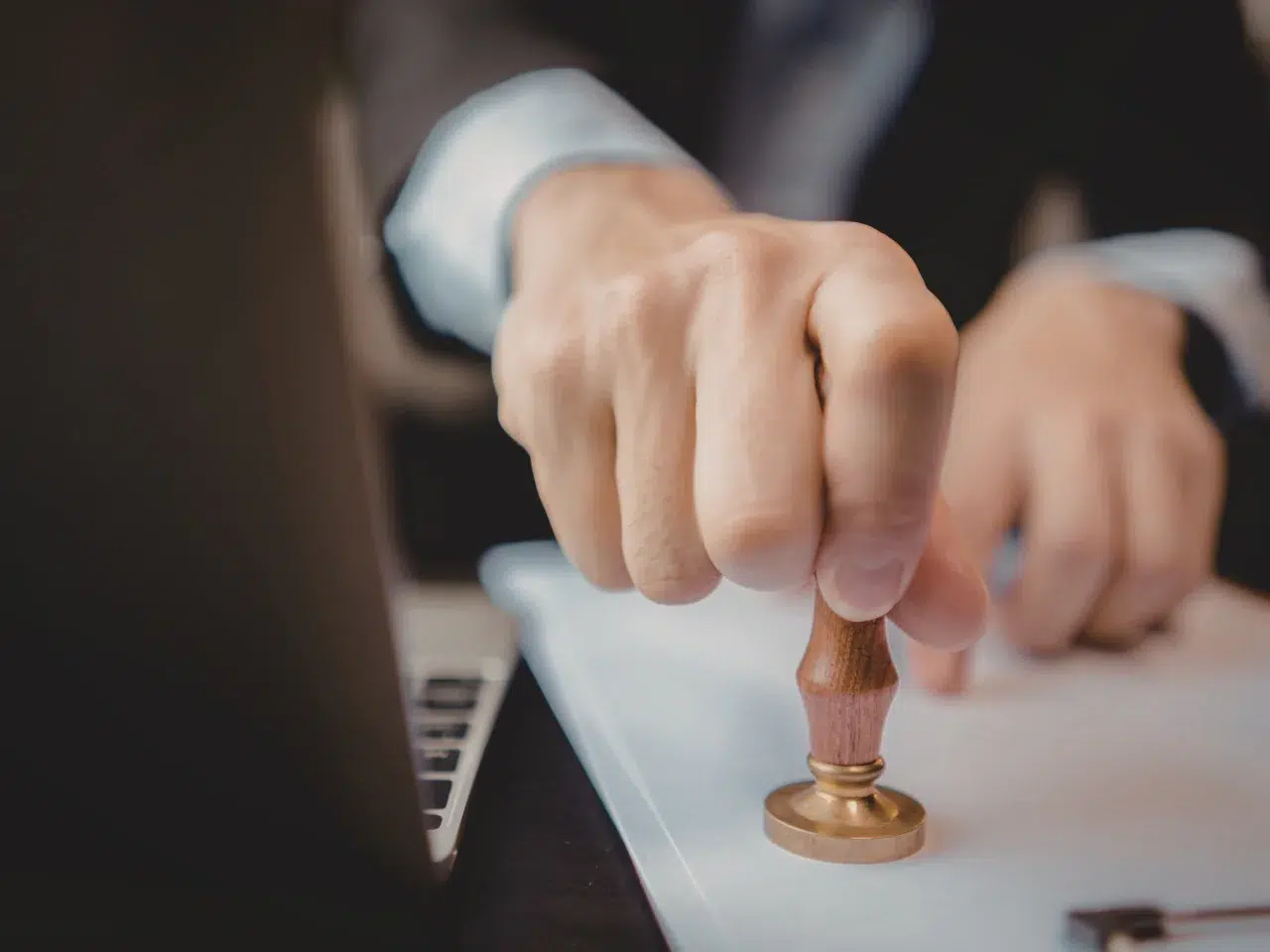 A close-up of a person’s hand pressing a wooden stamp onto a document, with part of a laptop visible on the desk.