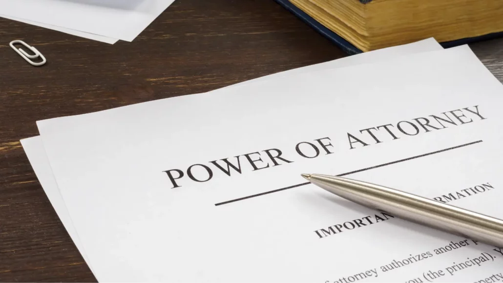 A close-up of a document titled Power of Attorney on a desk. A silver pen rests on the paper, and a thick book and paperclip are visible nearby.
