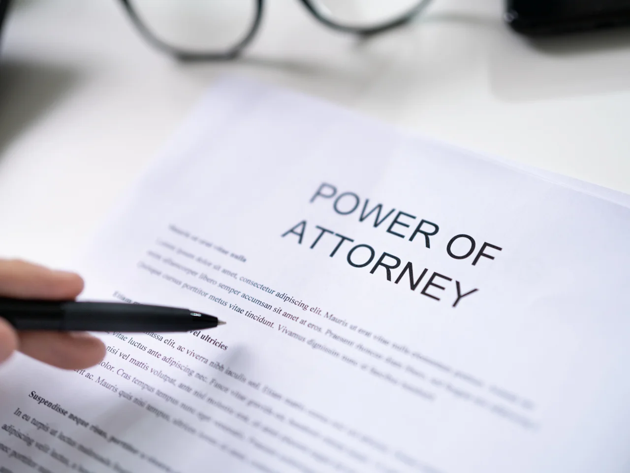 A hand holding a pen points to a document titled Power of Attorney on a desk, with a pair of eyeglasses placed in the background.