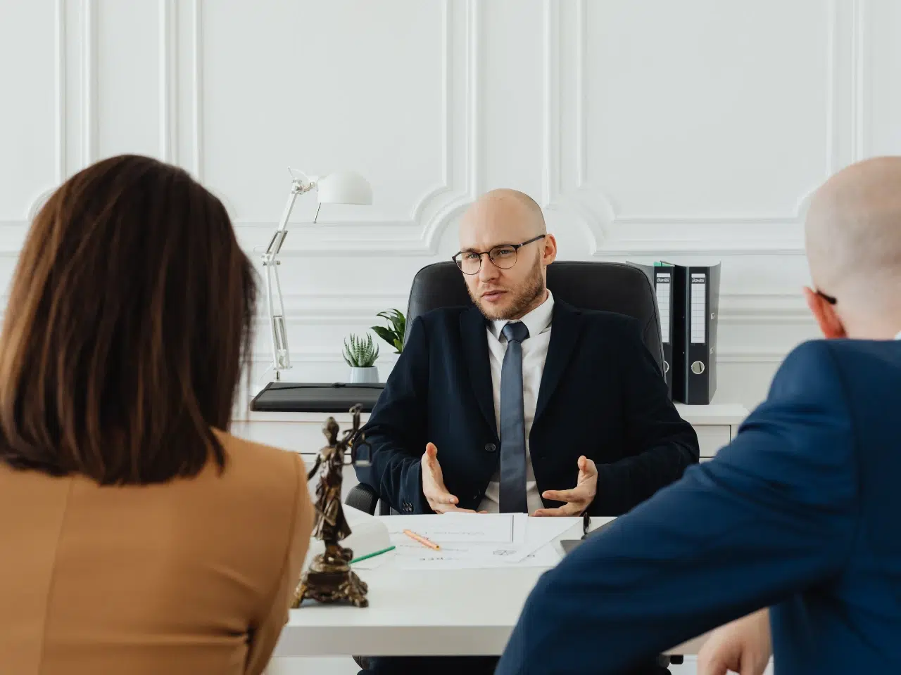 A man in a suit sits at a desk, speaking to two people across from him. Legal documents, a statue of Lady Justice, and office supplies are on the desk in a modern, white office setting.