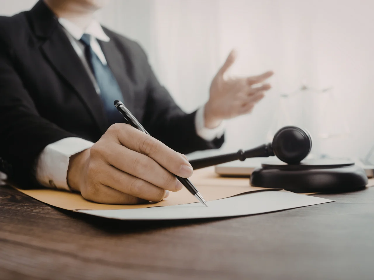 A person in a suit sits at a desk, holding a pen and writing on paper. A gavel and legal documents are on the table, suggesting a legal or judicial setting.
