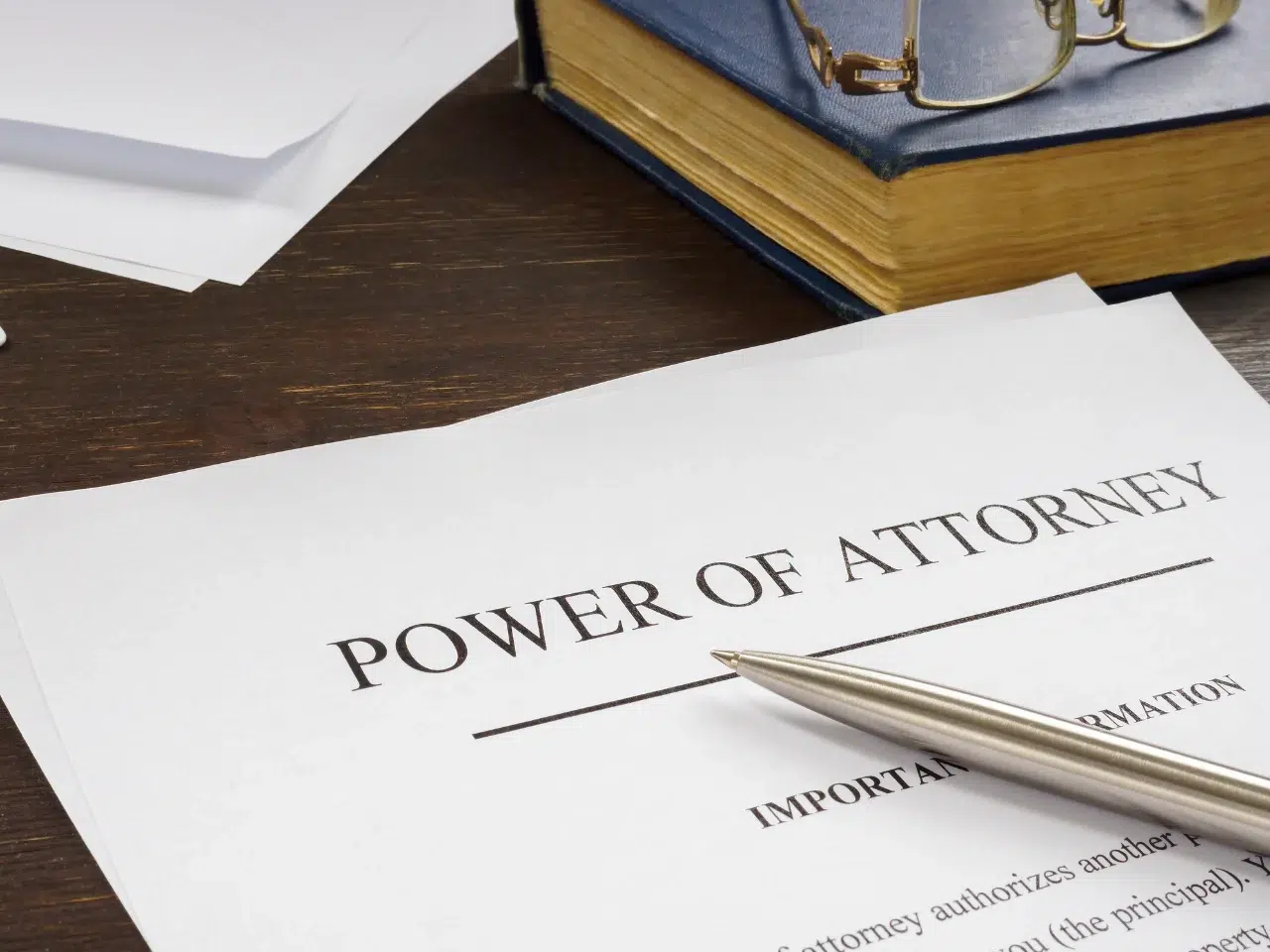 A document titled Power of Attorney sits on a desk next to a silver pen, a closed book with glasses on top, and a few loose papers in the background.