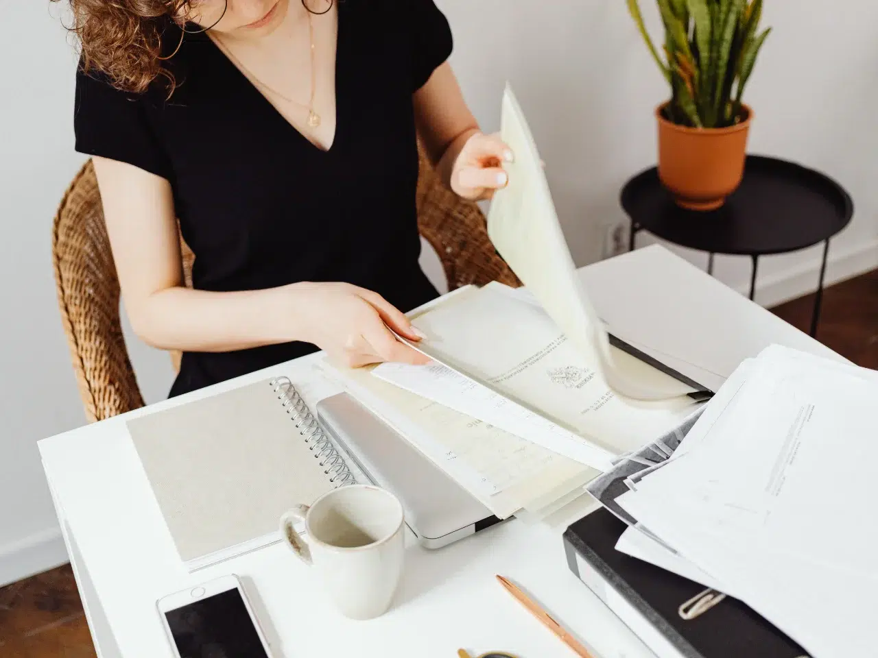 A woman in a black shirt sits at a desk looking through papers and documents. The desk is cluttered with notebooks, a coffee mug, a smartphone, and office supplies. A potted plant is in the background.