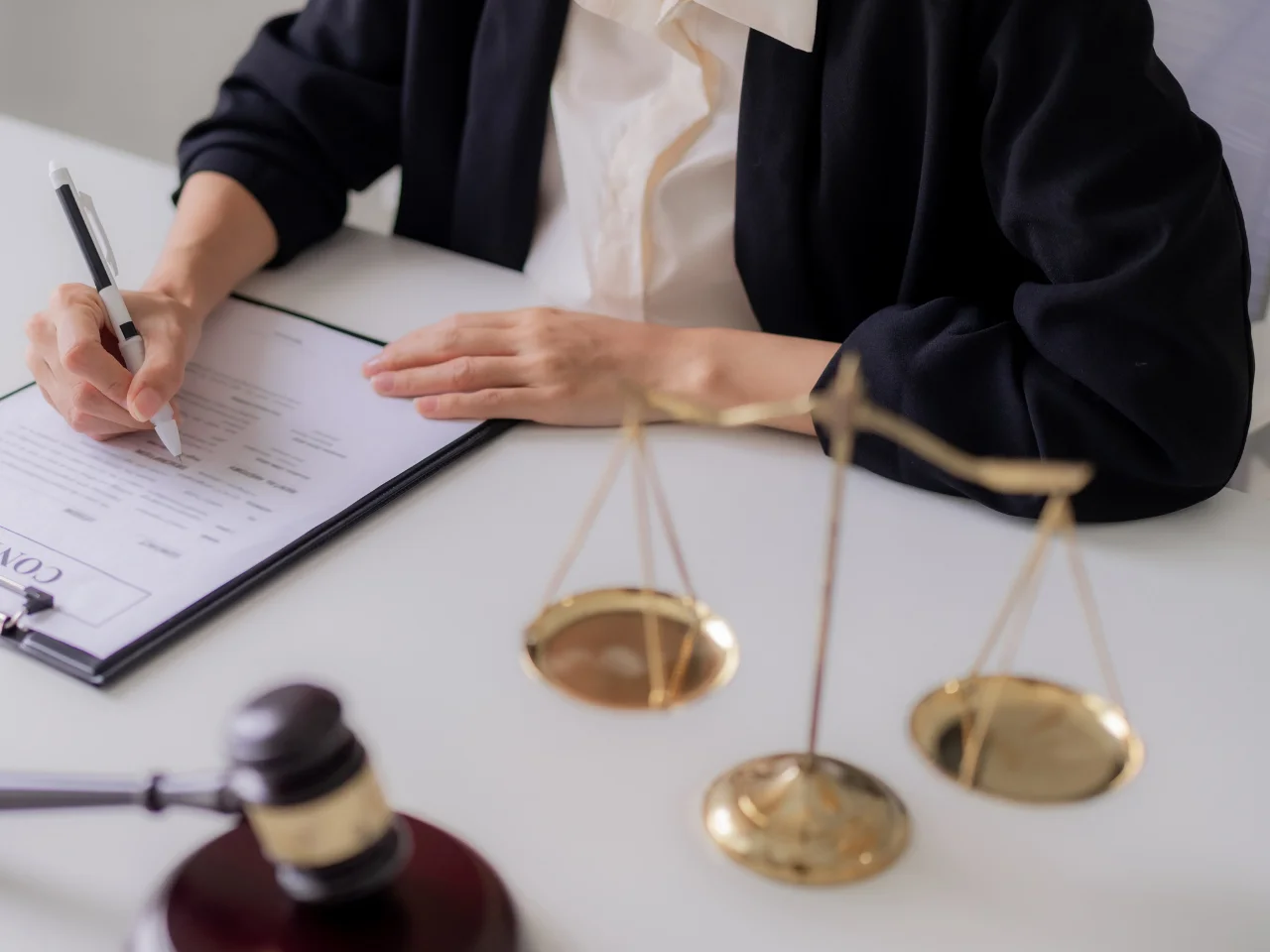 A person in professional attire fills out a document on a desk with a gavel and scales of justice in the foreground, symbolizing a legal or courthouse setting.