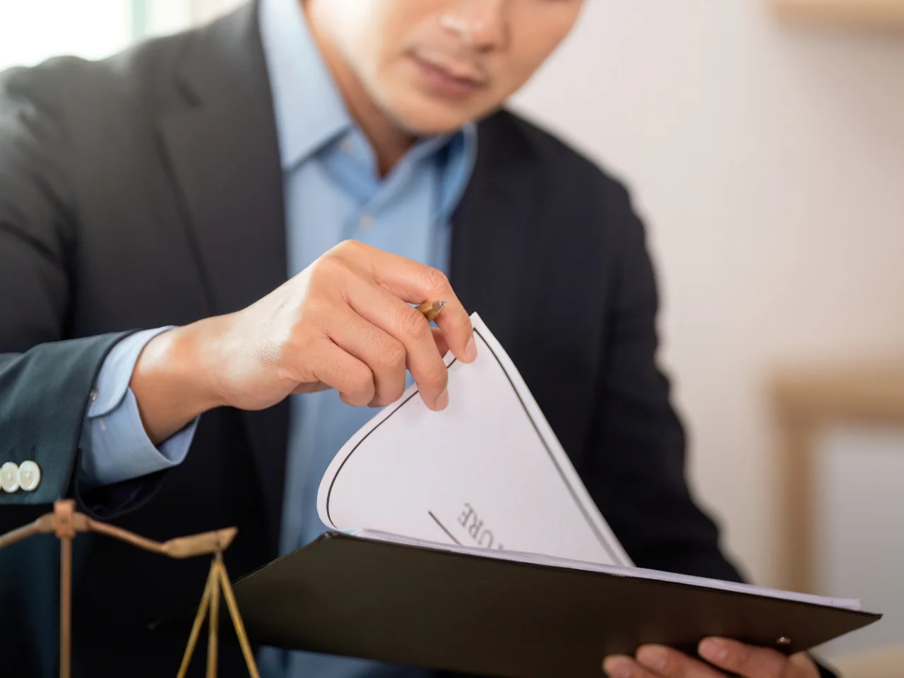 A person in a suit flips through documents on a clipboard, with a scale of justice visible on the desk, suggesting a legal or professional setting.