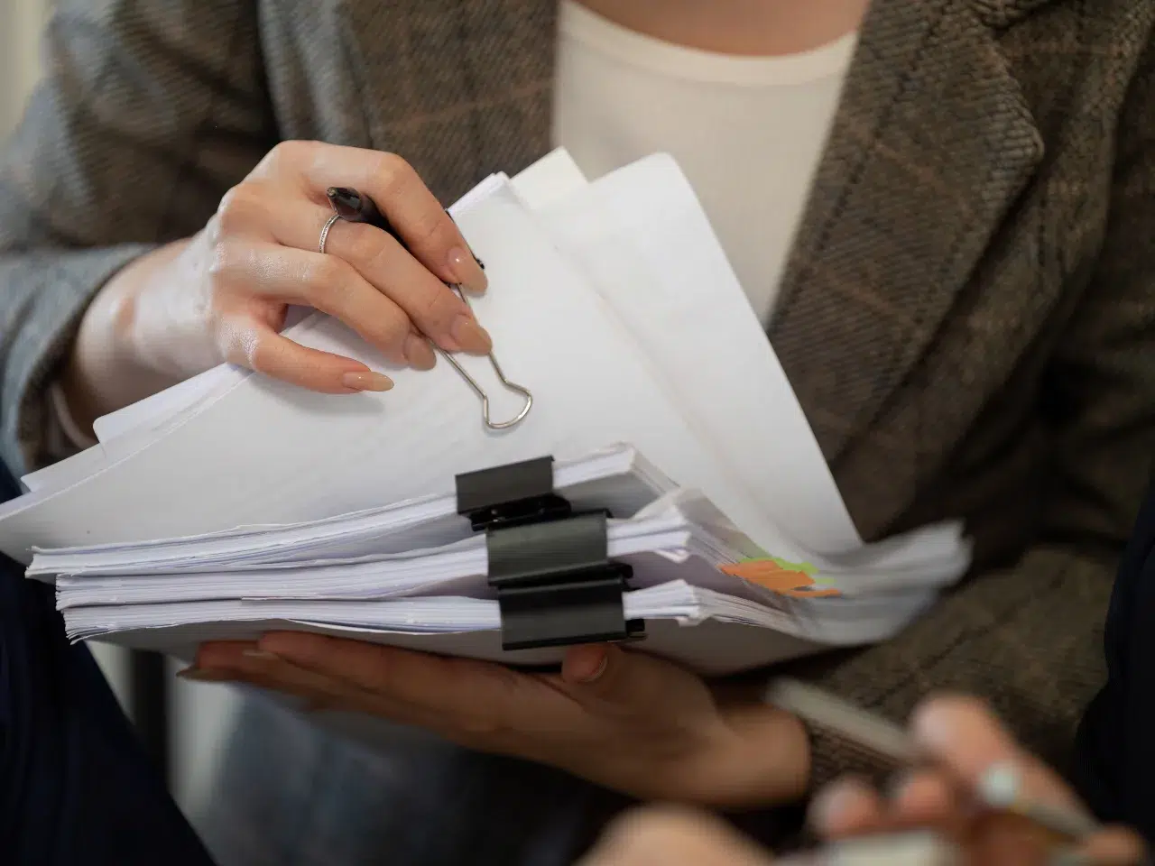 A person wearing a brown blazer holds a stack of documents clipped with binder clips, while holding a pen and preparing to write or review the papers.