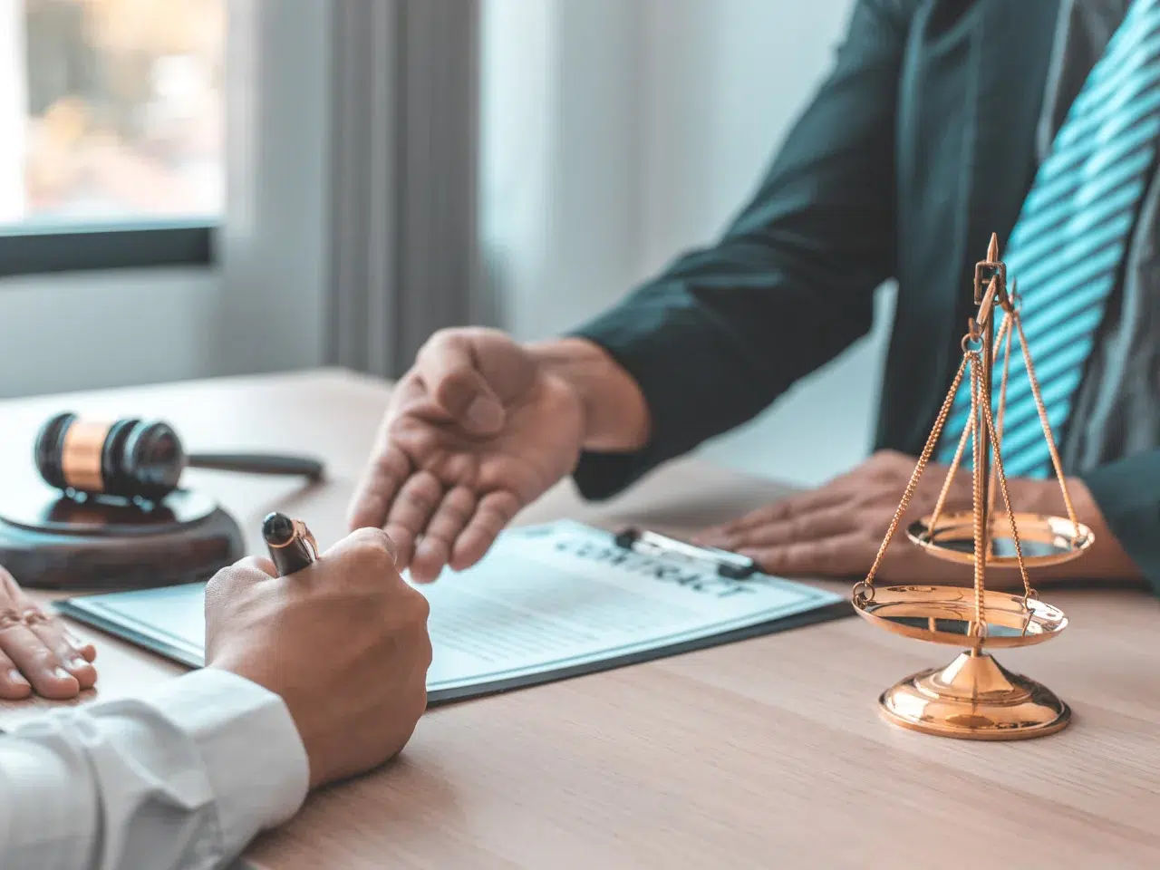 A person signs a document on a desk beside a gavel and a golden scale, with another person in a suit gesturing towards the paperwork, suggesting a legal or consultation setting.