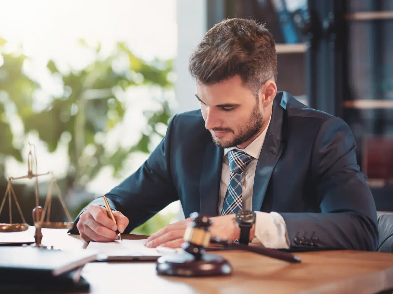 A man in a suit sits at a desk, writing on paper. A gavel, legal books, and scales of justice are on the desk, suggesting a legal or lawyer setting. Bookshelves and plants are visible in the background.