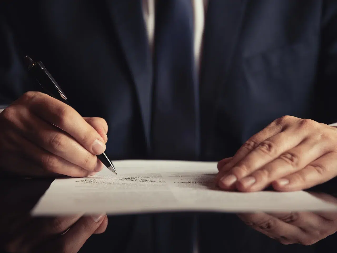 A person in a dark suit signs a document with a pen at a reflective desk, with both hands visible, focusing on the act of signing paperwork.
