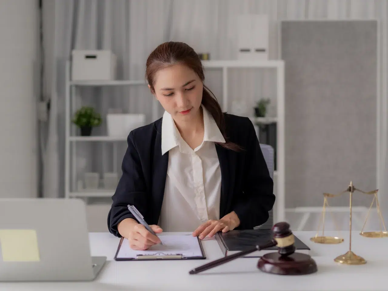 A woman in a suit sits at a desk, writing on a clipboard with a gavel, scales of justice, and a laptop nearby, in a bright office setting.